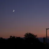 A tranquil crescent moon illuminates the evening sky over Caen, France, with silhouetted trees and a warm sunset backdrop.