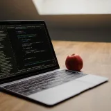 Open laptop displaying code next to a red apple on a wooden desk.