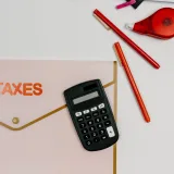 Imagen cenital de una calculadora, bolígrafos y una carpeta con la etiqueta «IMPUESTOS», ideal para temas relacionados con las finanzas.