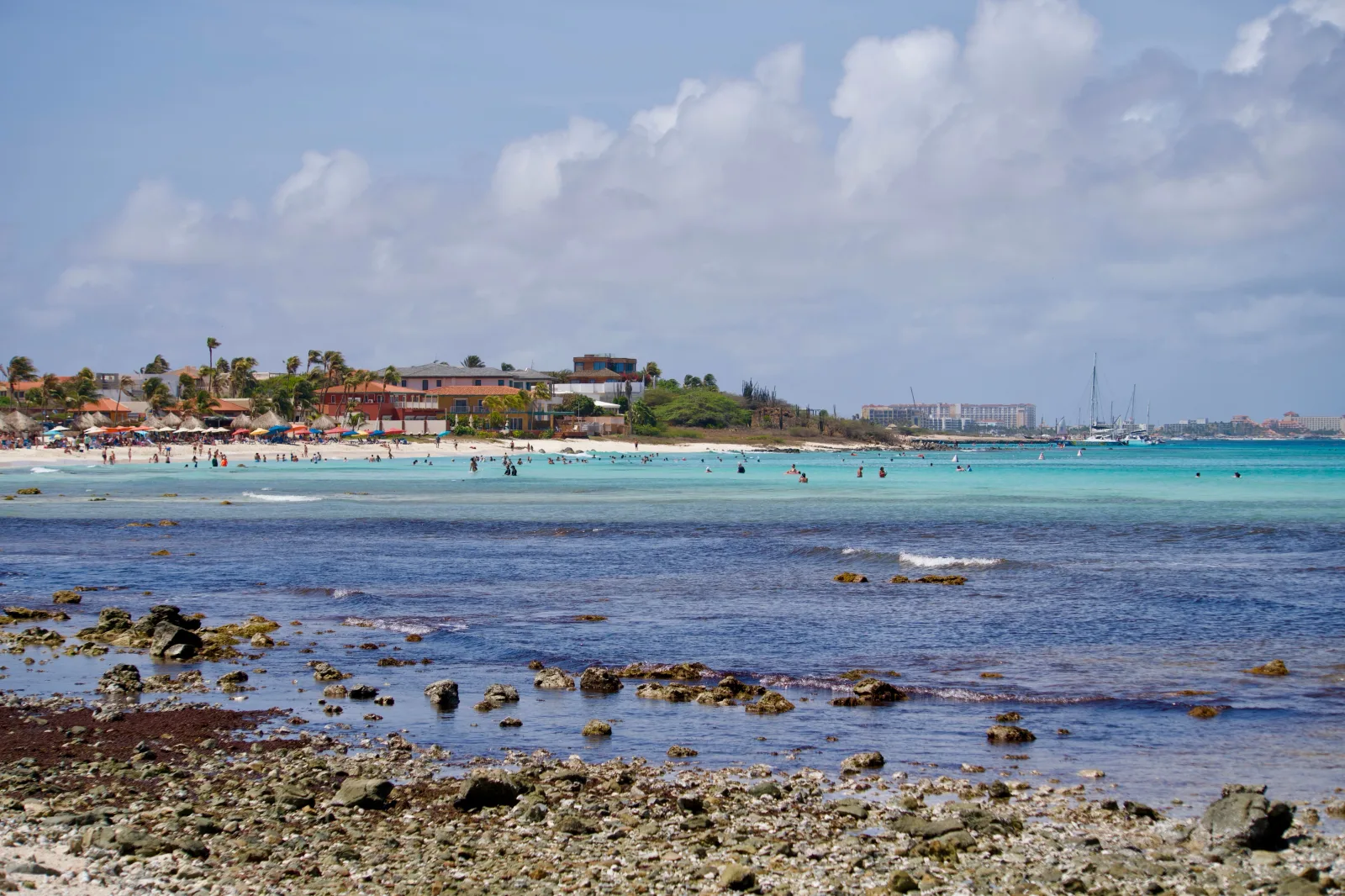 Una pintoresca escena de playa en Aruba, con aguas turquesas, una costa rocosa y animadas actividades playeras.