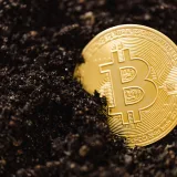 Primer plano de una moneda dorada de Bitcoin parcialmente enterrada en el suelo, que ilustra el concepto de minería de moneda digital.