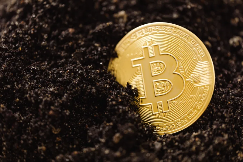Primer plano de una moneda dorada de Bitcoin parcialmente enterrada en el suelo, que ilustra el concepto de minería de moneda digital.