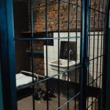 Interior de una celda de prisión con poca luz, con una cama, un lavamanos y paredes de ladrillo expuestas, que evoca sentimientos de aislamiento.