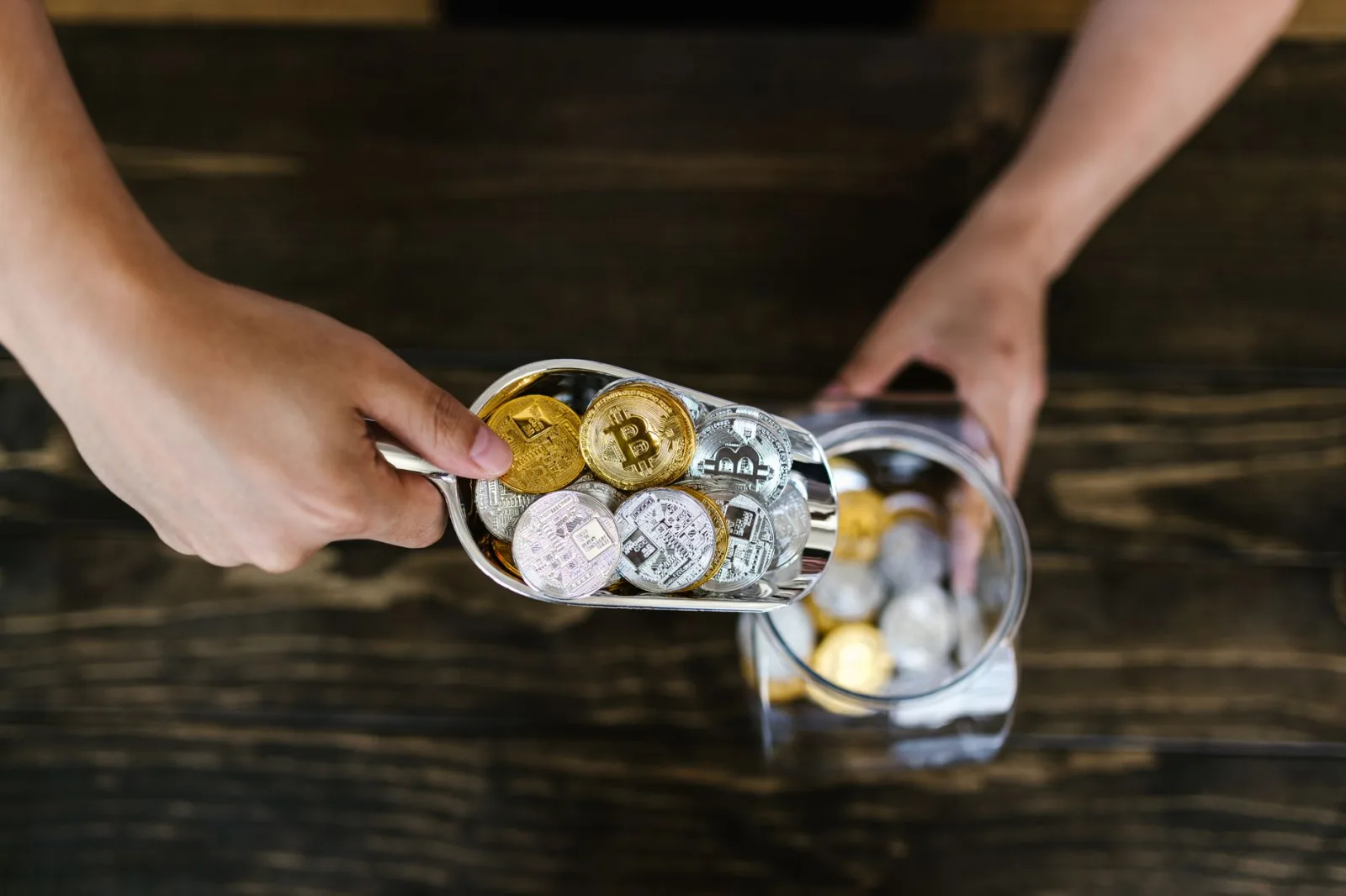 Primer plano de unas manos recogiendo varias monedas de criptomonedas y metiéndolas en un frasco sobre una superficie de madera.