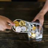 Primer plano de unas manos recogiendo varias monedas de criptomonedas y metiéndolas en un frasco sobre una superficie de madera.