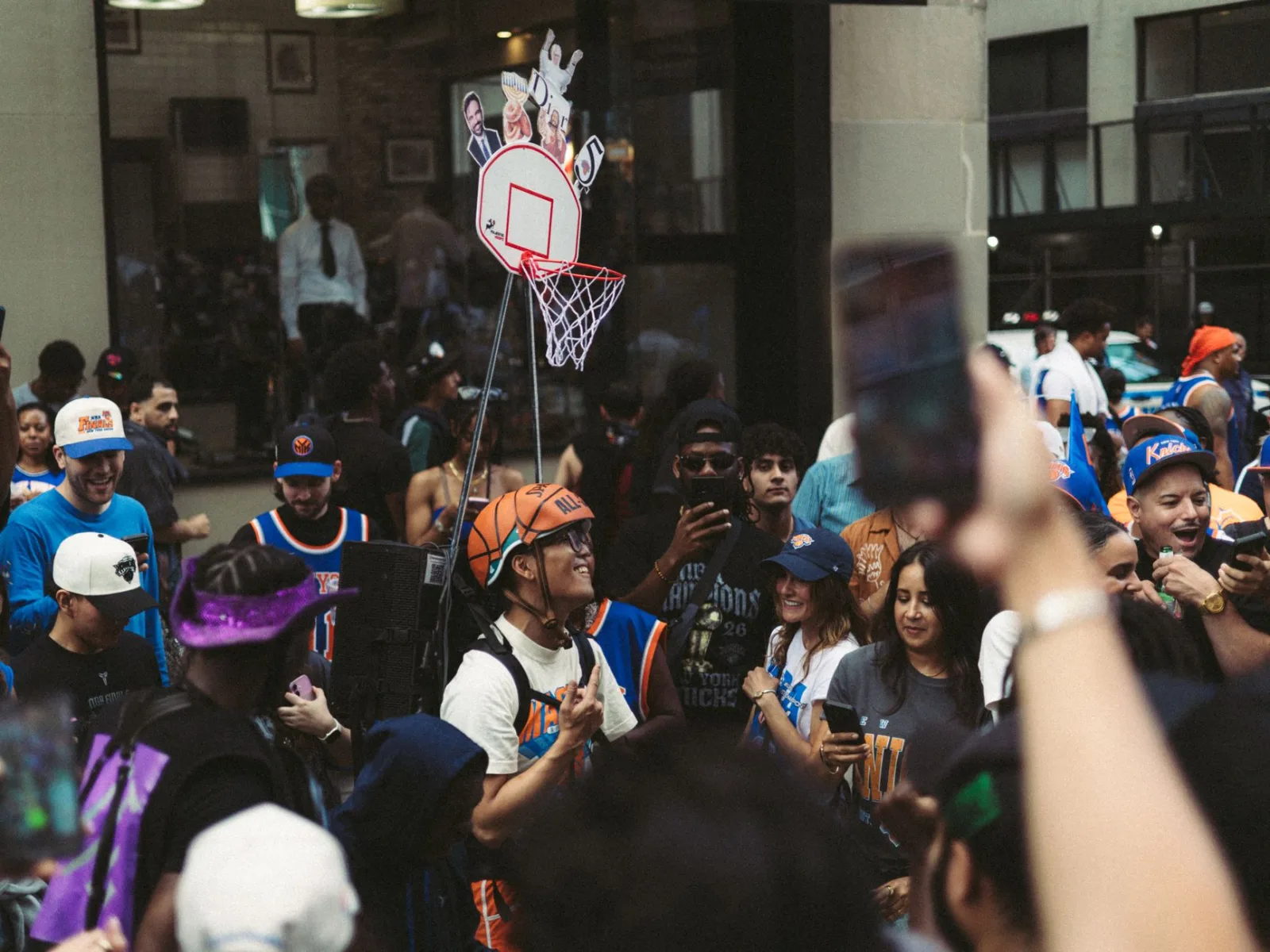 Animada escena callejera de aficionados al baloncesto que llevan camisetas y celebran junto a una canasta pequeña.